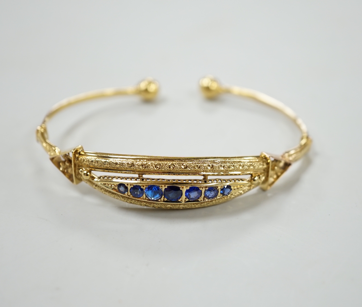 A 750 yellow metal and graduated seven stone sapphire?, paste and doublet set open work bangle, gross weight 12.1 grams.